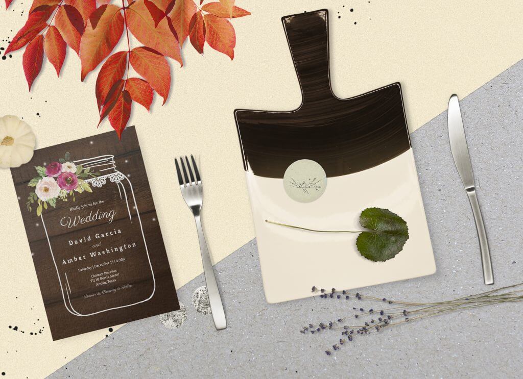 Rustic Mason Jar Wedding Invitation & Dinner Plate Set. The invite showcases a charming line drawing of a mason jar with colorful flower illustration. Text is centered. Background is wooden. Nearby are a cutting board, fork, and knife.