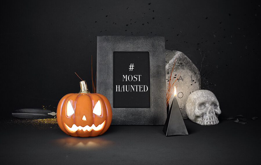 Stone photo frame with a card inside reading 'Most Wanted'. Set against a dark background, with a skull nearby, alongside a tombstone and a illuminated carved pumpkin.