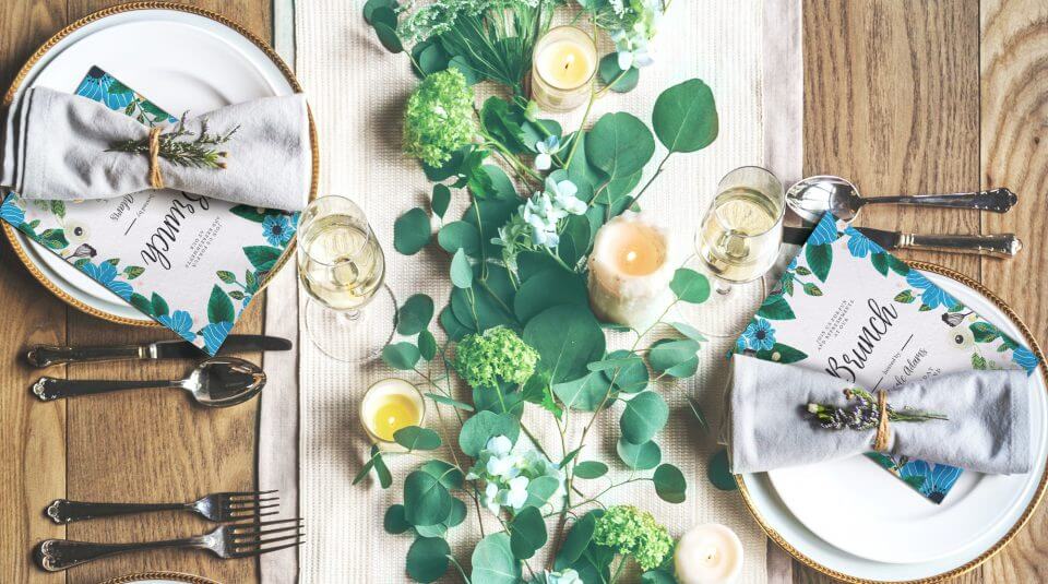 Elegant Brunch Table Setting: Invitations featuring delicate blue flower designs peek from beneath folded napkins atop each plate. The table is adorned with vibrant fresh blooms and verdant leaves