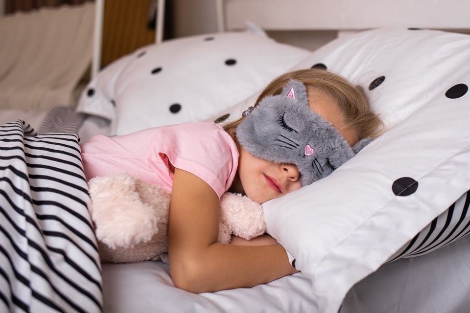 1-little-girl-sleep-mask-sleeps-bed-with-stuffed-toy (1) Young girl asleep in bed wearing a gray cat sleep mask, cuddling a stuffed toy, surrounded by cozy pillows and blankets, capturing a calm sleepover or bedtime moment.