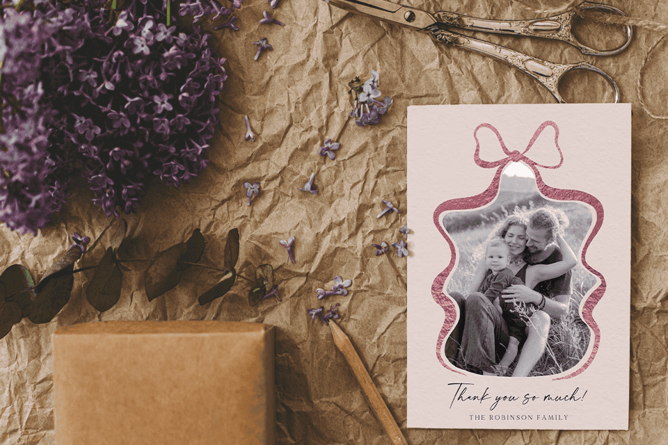 1 thank-you-card-housewarming-card-bow-family-photo (2) Rustic flat lay featuring a housewarming thank-you card with a family photo framed by a pink bow illustration, surrounded by kraft paper, lilac flowers, vintage scissors, and gift-wrapping elements.