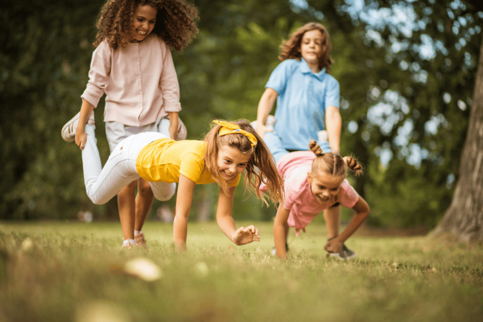 birthday party outdoor games (1) Group of children laughing and playing an outdoor relay-style game on the grass, crawling and racing together in a sunny park during a kids’ birthday party.