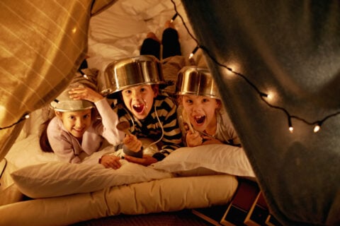 Three kids lying on pillows inside a cozy blanket fort, wearing metal pots as playful hats, laughing and cheering under warm string lights during a fun slumber party.