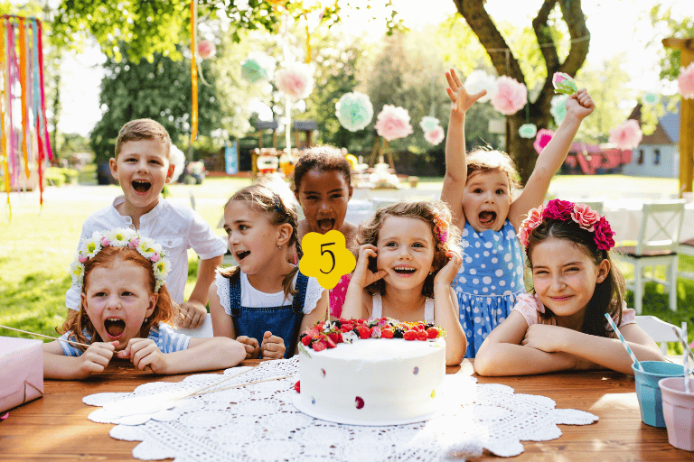 Group of excited kids gathered around a birthday cake with a yellow number 5 candle, cheering at an outdoor garden birthday party decorated with pastel hanging pom-poms and ribbons.