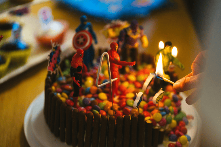 Close-up of a colorful superhero-themed birthday cake topped with candy pieces, small action figure characters, and a number four candle being lit by hand, set on a party table during a child’s birthday celebration.