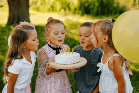 Four young girls outdoors blowing out candles on a birthday cake, holding the cake together on a wooden platter, surrounded by greenery and a yellow balloon, capturing a joyful kids’ birthday celebration.