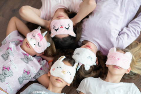 Top-down view of five children lying in a circle on the floor at a slumber party, wearing cute animal sleep masks and cozy pajamas, suggesting a fun, relaxed sleepover atmosphere.