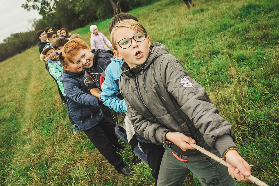 boy-birthday-kids-activities-outdoor Group of children playing an outdoor tug-of-war game on a grassy field, with boys in jackets pulling a rope together, showing energetic teamwork during a kids’ party or group activity.
