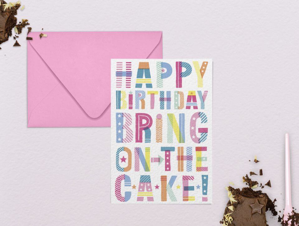 Vibrant Typography Birthday Card: 'Happy Birthday, Bring on the Cake' with Diverse Colors and Patterns. Set on a Soft Pink Surface Alongside a Closed Pink Envelope and a Tempting Slice of Cake.