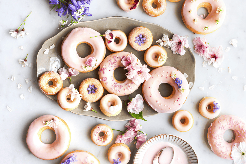 donuts sleepover party food (1) Assorted pink-glazed donuts and mini doughnuts arranged on a neutral platter, decorated with edible flowers, pastel sprinkles, and scattered blossoms, creating a soft, whimsical dessert spread perfect for a sleepover or party.