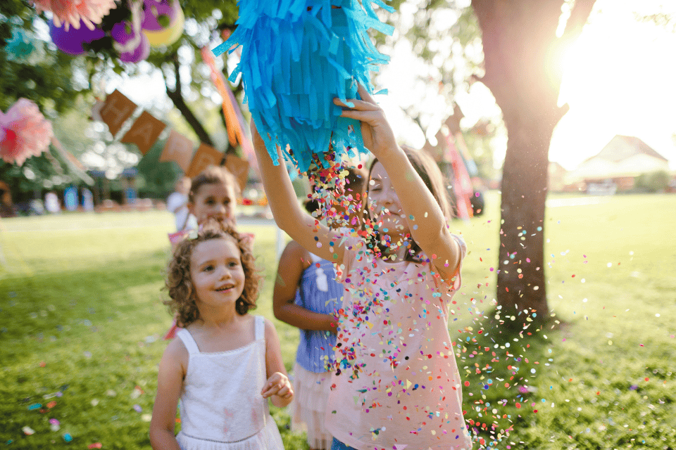 girl birthday celebration party (1) Children outdoors at a birthday party as a girl lifts a blue piñata and colorful confetti rains down, with other kids watching excitedly in a sunny park decorated with balloons and party banners.