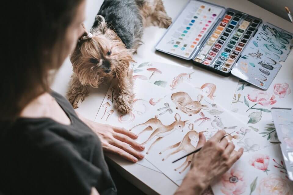 Designer's studio scene with watercolor and sketched animal designs; the artist is engrossed in drawing, with a vibrant watercolor palette on the table, and her small dog sitting loyally by her side.