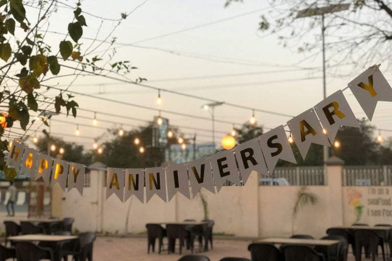 Outdoor anniversary celebration with a “Happy Anniversary” banner strung across a patio, surrounded by string lights and seating.