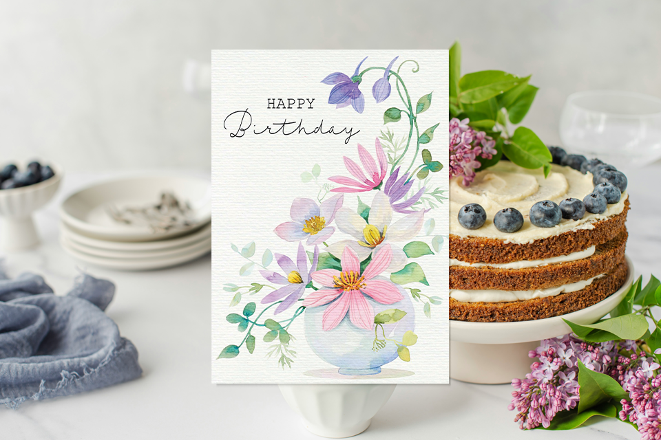 happy-birthday-boss-card-floral Elegant floral birthday card with watercolor flowers in a vase and the words “Happy Birthday,” displayed beside a layered cake topped with blueberries and lilac flowers on a bright, minimal table setting.
