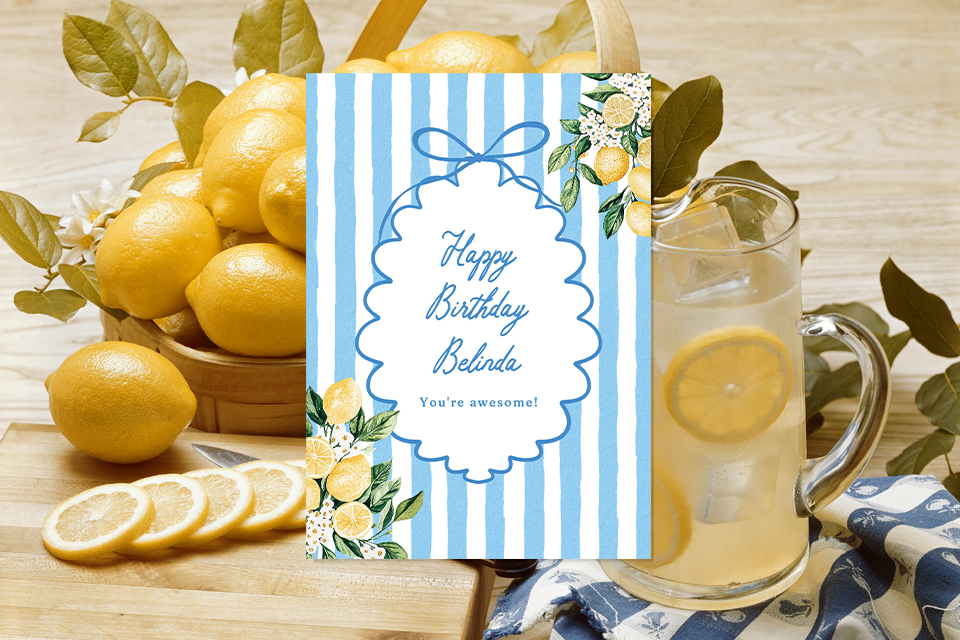 happy-birthday-card-stripes-lemons Blue-and-white striped birthday card with lemon illustrations and a ribbon frame reading “Happy Birthday Belinda” placed on a wooden table with fresh lemons and a glass pitcher of lemonade.