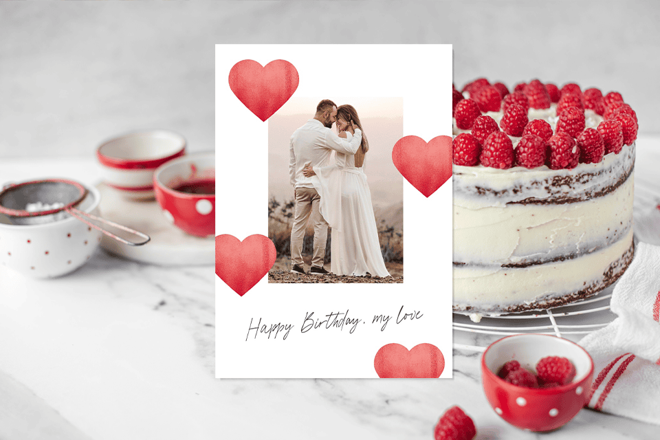 happy-birthday-card-wife-photo-hearts (1) Romantic birthday card featuring a couple embracing in a photo framed by red heart illustrations, with the message “Happy Birthday, my love,” styled against a dessert scene with a raspberry cake and red bowls.