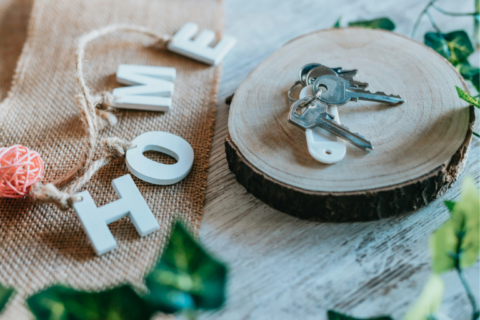 Rustic housewarming scene with silver house keys resting on a round wooden slice, next to burlap fabric and decorative letters spelling “HOME” on a light wooden surface with greenery accents.