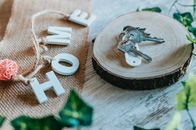 Rustic housewarming scene with silver house keys resting on a round wooden slice, next to burlap fabric and decorative letters spelling “HOME” on a light wooden surface with greenery accents.