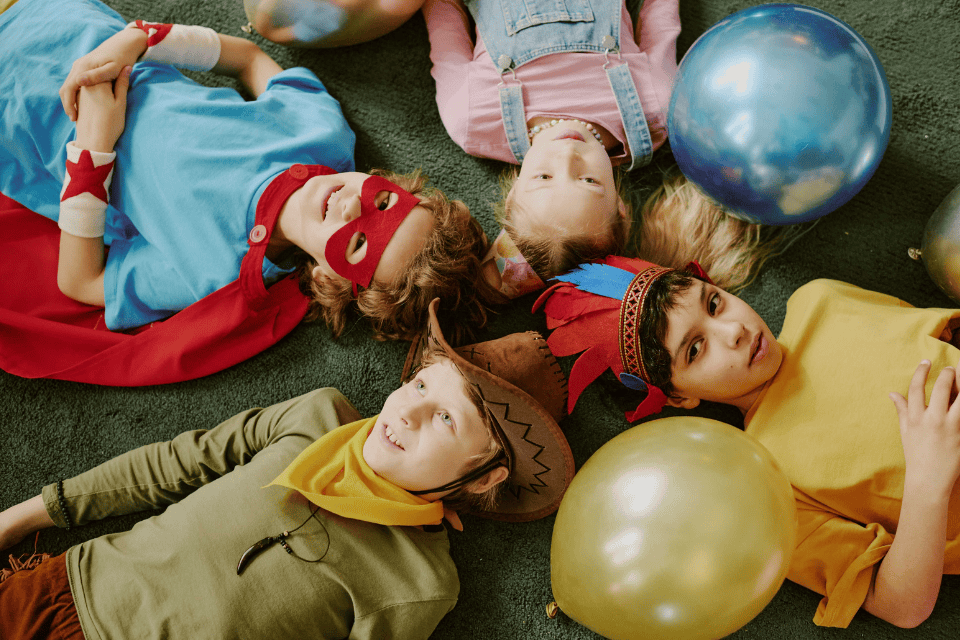 kids birthday decorations kids in costumes (1) Children in playful costumes lying in a circle on a carpet during a birthday party, surrounded by colorful balloons, smiling and looking up at the camera.