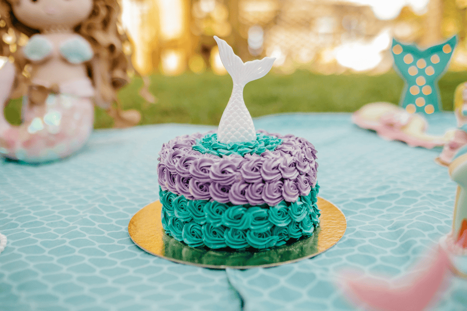 kids birthday party cake (1) Mermaid-themed kids’ birthday cake with teal and lavender rosette frosting, topped with a white mermaid tail, displayed on a gold cake board on a turquoise party table outdoors.