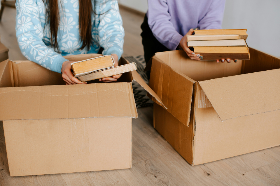 new house unpacking books cardboard boxes Two people unpacking books from open cardboard boxes on a wooden floor, placing stacked hardback books into boxes while settling into a new home.