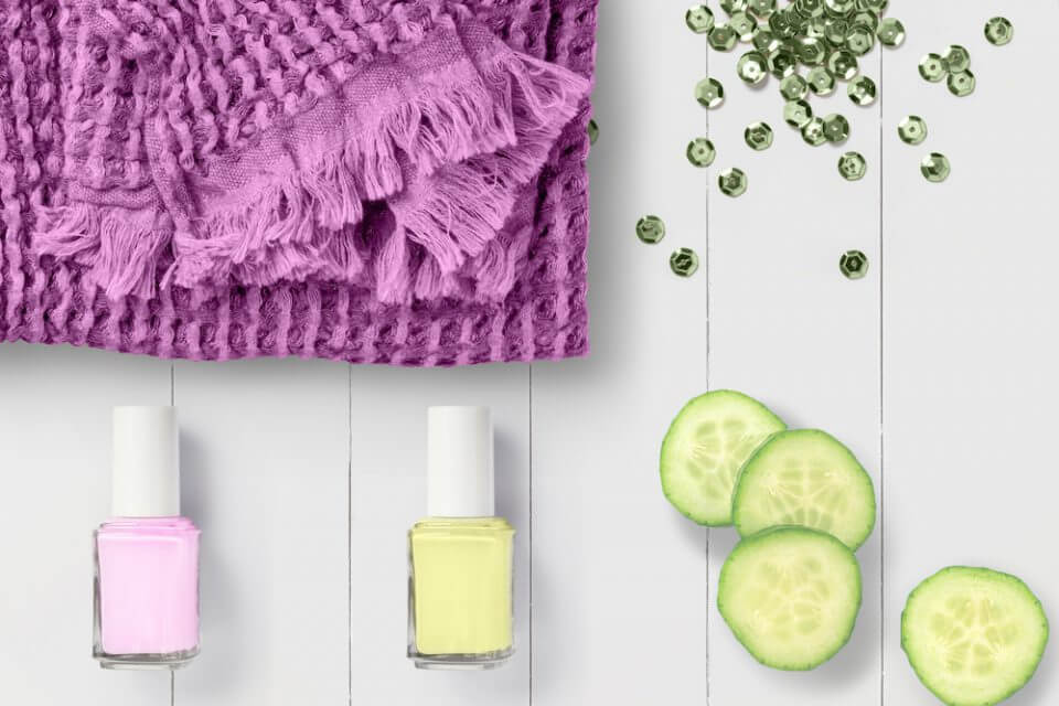 From an overhead view, a spa party setup featuring two bottles of nail polish (one in soft pink, the other in light yellow), a pink towel, and refreshing cucumber slices for a pampering experience.