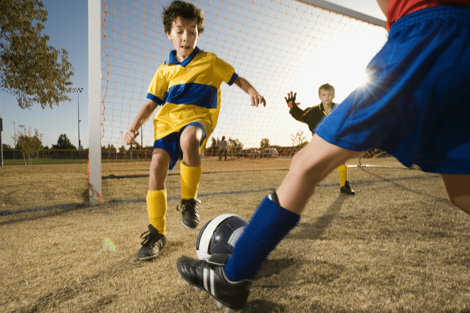 sports birthday ideas boys tournament soccer (1) Kids playing a soccer match on a sunny field, with a boy in a yellow and blue jersey kicking the ball near the goal while another player defends and a young goalkeeper watches, capturing an energetic youth sports moment.