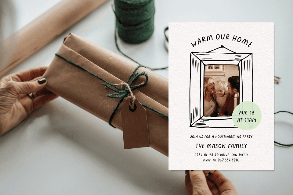 warm-our-house-housewarming-invite-photo (1) Hands holding a rustic housewarming invitation beside a gift wrapped in brown paper and green twine, featuring a family photo inside a hand-drawn frame with the text “Warm Our Home.”