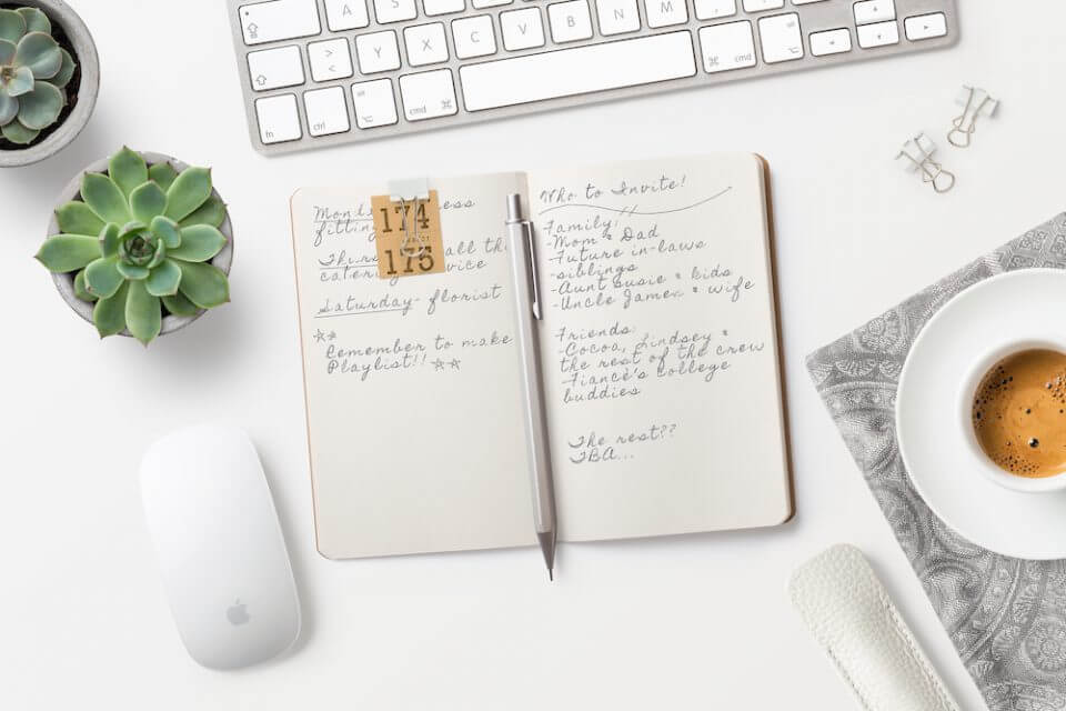 Wedding Guest List Planning: Handwritten Notes on Open Notepad with Pencil, Surrounded by Office Supplies and Coffee on White Surface.