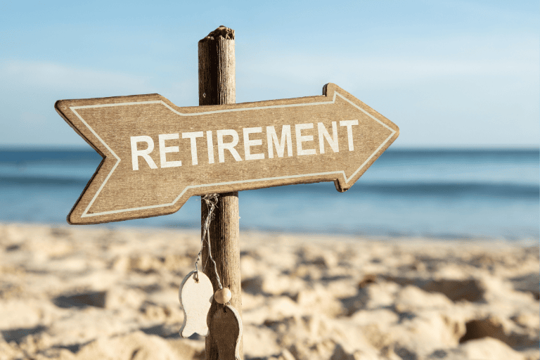 Wooden arrow sign reading ‘Retirement’ pointing right, standing on a sandy beach with the ocean and clear blue sky in the background.