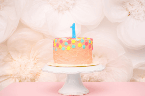 Pastel pink first birthday cake with colorful polka dots and a blue number one candle on a white cake stand, styled against a soft floral backdrop.