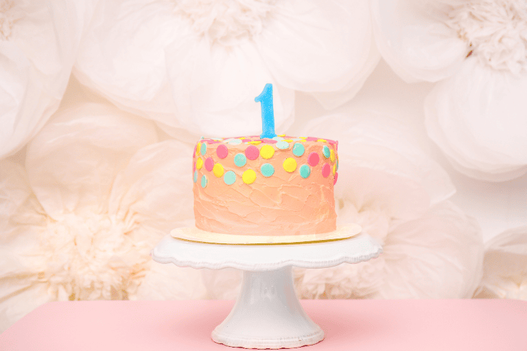 Pastel pink first birthday cake with colorful polka dots and a blue number one candle on a white cake stand, styled against a soft floral backdrop.