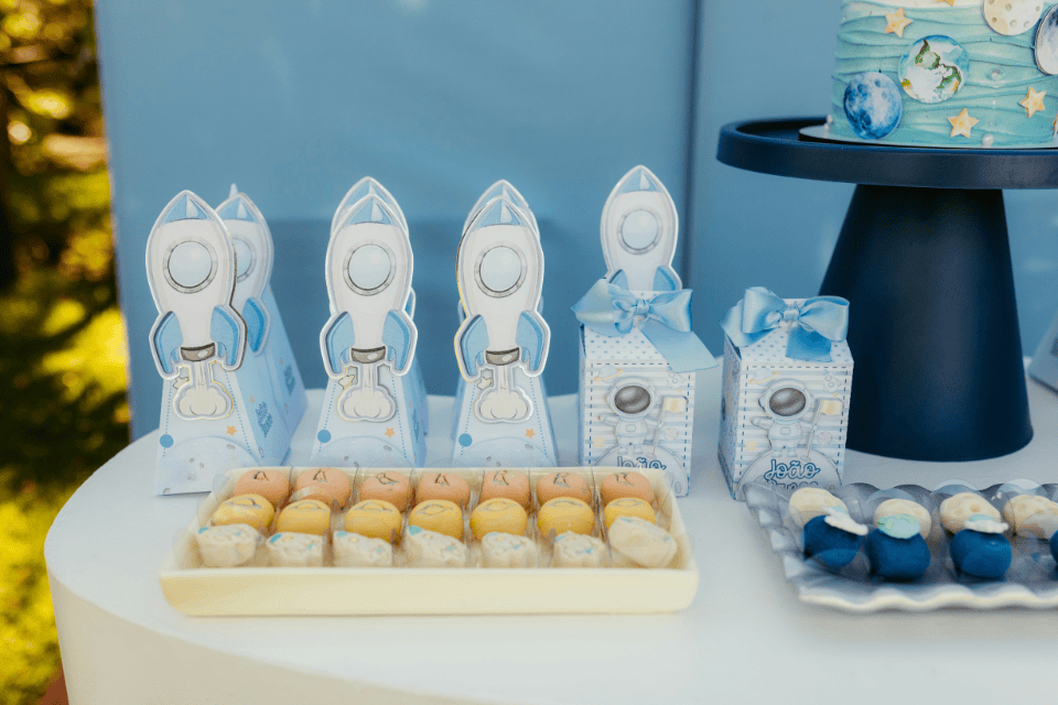 1st birthday theme decor Space-themed first birthday dessert table with blue and white rocket-shaped favor boxes, astronaut gift boxes tied with blue bows, assorted bite-size sweets, and a matching planet-decorated cake on a stand, styled in soft pastel blue tones outdoors.