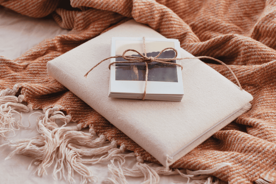 3 retirement party ideas notes memories (1) Neutral-toned retirement keepsake arrangement with a linen-covered book, a stack of printed photos tied with twine, and a cozy textured throw, styled as memory notes or mementos for a retirement celebration