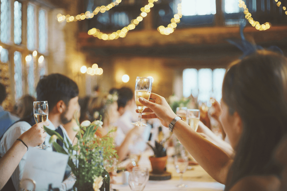 5 retirement party celbration toast (1) Guests raising champagne glasses in a warmly lit indoor venue with string lights, celebrating a retirement party at a long table filled with flowers, candles, and glassware, creating an elegant and joyful atmosphere.