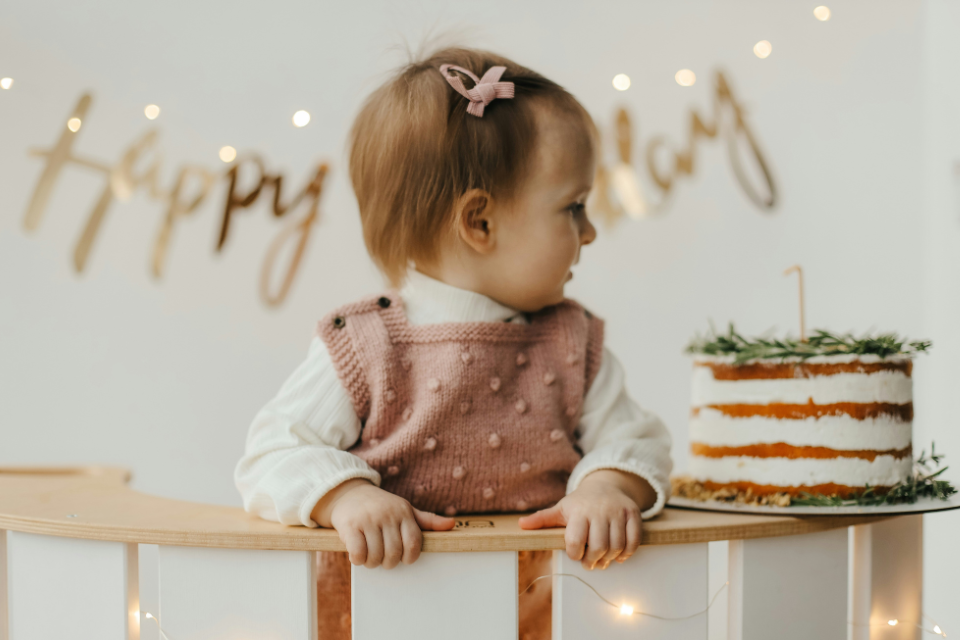 baby 1st birthday celebration Baby girl at a first birthday celebration wearing a pink knit outfit, standing at a small table beside a layered birthday cake with a single candle and soft party lights in the background.