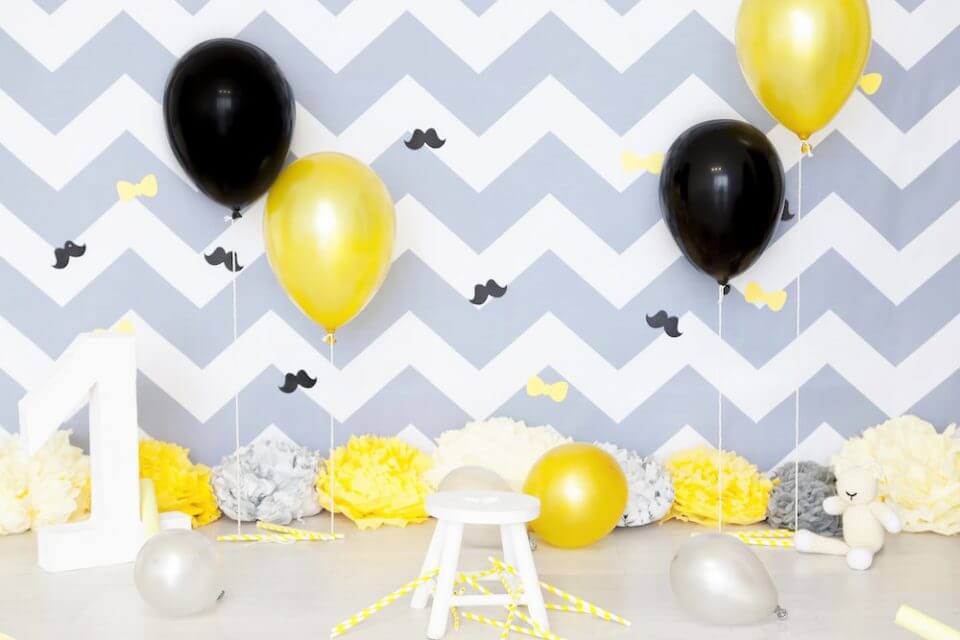 Birthday Decor: Grey, Yellow, Black, and White Balloons Against Zigzag Grey and White Backdrop. White 'One' Stands Out with Solo Bench.