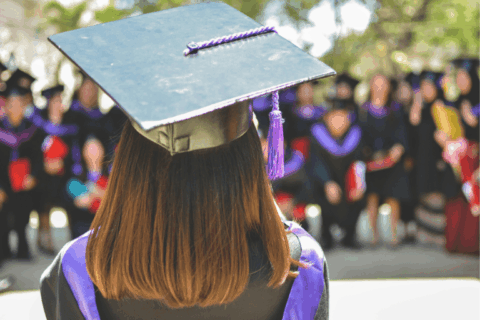 Graduate wearing a mortarboard and gown seen from behind, standing on stage during an outdoor graduation ceremony, with seated graduates blurred in the background.