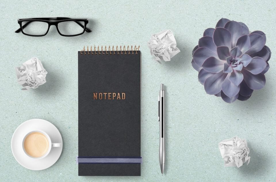 A notepad accompanied by a cup of coffee, a bouquet of flowers, eyeglasses, and a pen. This scene conveys a serene and creative workspace, where one can enjoy a beverage, gather thoughts, and jot down ideas.