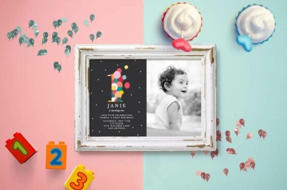 First birthday party planning guide: Vibrant invitation with '1' formed from balloons, set against a black backdrop with white dots. A cherished child's photo in the frame. Placed on a half light blue, half light pink background, with building blocks and cupcakes nearby