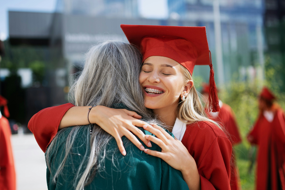 graduation ceremony family friends Smiling graduate in a red cap and gown hugging an older woman during a graduation ceremony, with other graduates blurred in the background, capturing an emotional family celebration moment.