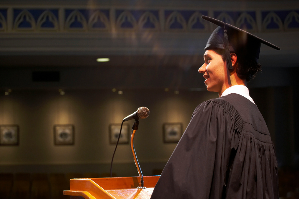 graduation ceremony woman speech Graduate in a cap and gown standing at a podium, delivering a speech into a microphone on a softly lit stage during a graduation ceremony.