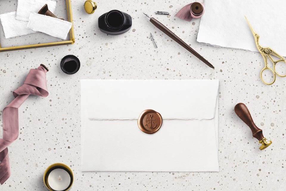 Flatlay of Wedding Invitation Envelope Sealed with Pink Wax on Marble Surface. Accompanied by Small Candle, Scissors, Pink Ribbon, and Black Ink.