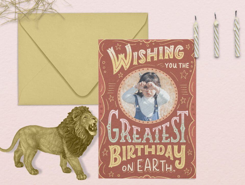 Universal Birthday Greetings & Heartfelt Card Message: Light Brown Card with a Festive Announcement 'Wishing You the Greatest Birthday on Earth', Featuring a Central Circus-like Photograph. Adjacent to a Playful Circus Lion Drawing and a Neatly Closed Envelope on a Pink Surface.