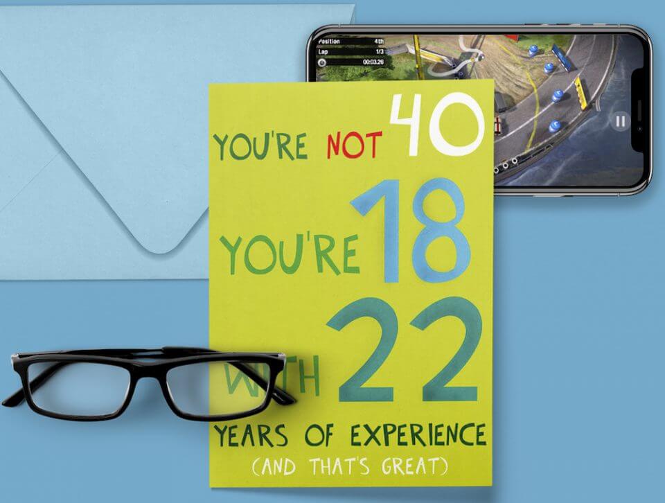 Humorous Birthday Greetings: 'You're Not 40, You're 18 with 22 Years of Experience, and That's Great!' on a Green Card. Positioned Against a Blue Background with Eyeglasses, a Smartphone, and a Sealed Envelope Nearby.