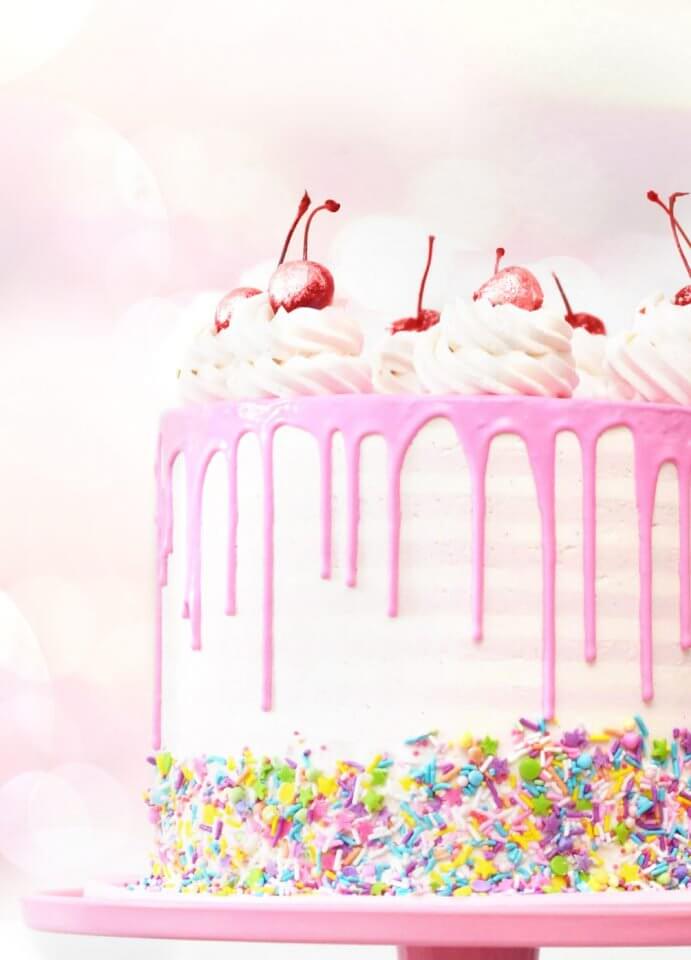 Vibrant cake adorned with colorful sprinkles, pink dripping icing, and luscious cherries on top, photographed from upclose