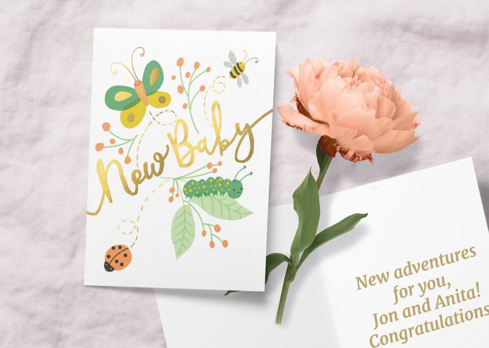 New Baby message in radiant gold, complemented by an enchanting illustration of flowers, butterfly, bee, and ladybug in shades of green and yellow. The card rests beside a delicate bloom and an open card bearing warm greetings.