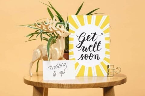 "A blog post cover for 'Best Get Well Wishes & Quotes For A Speedy Recovery', displaying an artfully designed 'Get Well Soon' greeting card. The card, prominently placed on a wooden tabletop, features an illustration of cheerful sunrays. Beside the card, there's a vase of elegant flowers, adding a touch of beauty and warmth to the scene, symbolizing care and recovery.