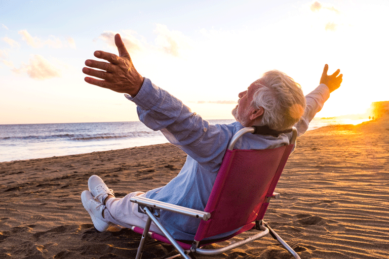 Senior man relaxing on beach chair at sunset with arms wide open, enjoying peaceful summer vacation by the ocean.