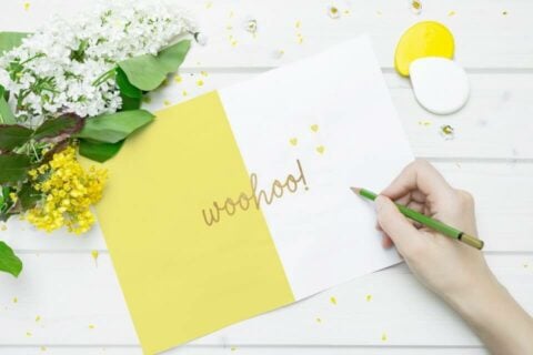 50 Congratulations Wishes & Quotes for Cards: Celebrating Achievements – With a Handwritten 'Woohoo' on a Cheerful Yellow and White Card, Accompanied by Flowers Nearby.
